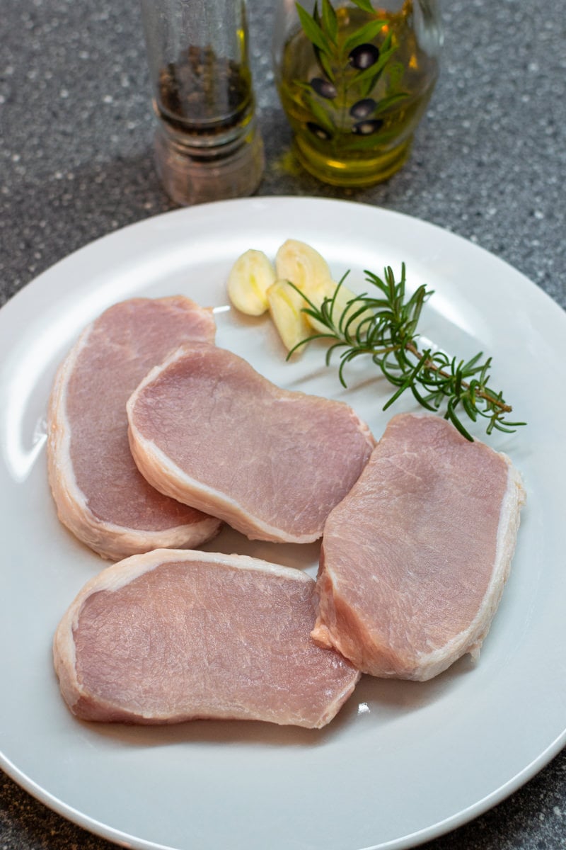 4 Pieces of pork chops with rosemary and garlic cloves on a white plate.