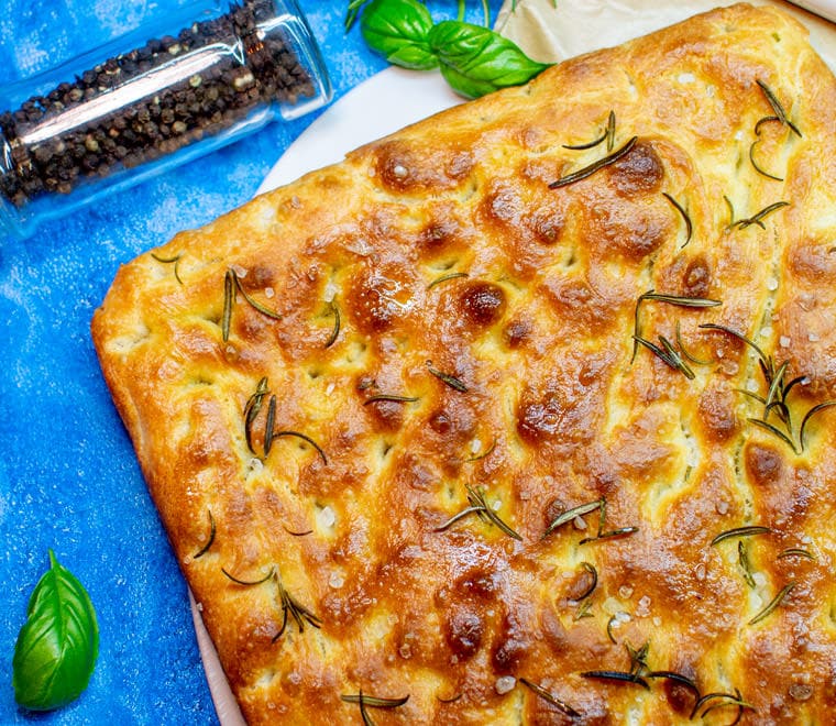 Classic Focaccia with rosemary on a blue background with black pepper.