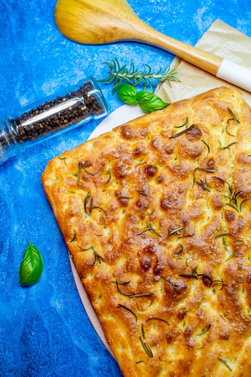 Classic Focaccia on a blue table with rosemary and black pepper.