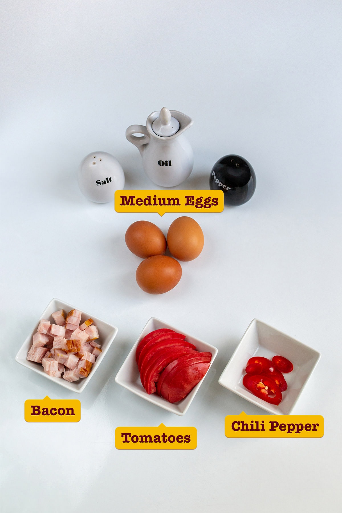 Eggs, cubes of bacon, slices of tomatoes and spicy pepper on white table.