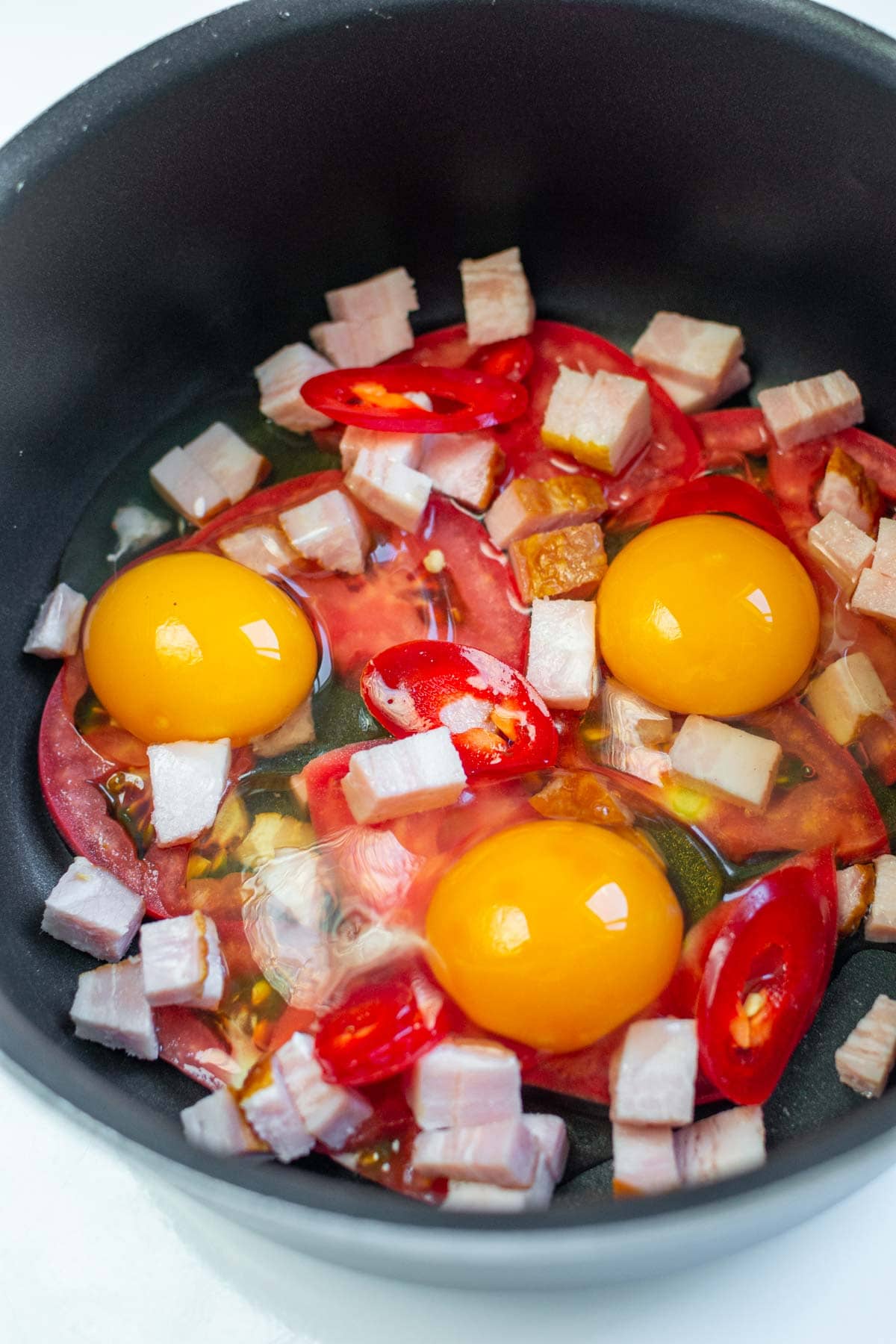Cubes of bacon, 3 raw eggs, tomatoes and spicy pepper in a pan.