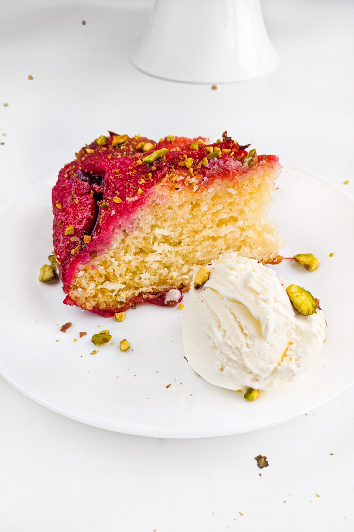 Slice of yogurt plum cake with crushed pistachios and ice cream ball on a white plate.