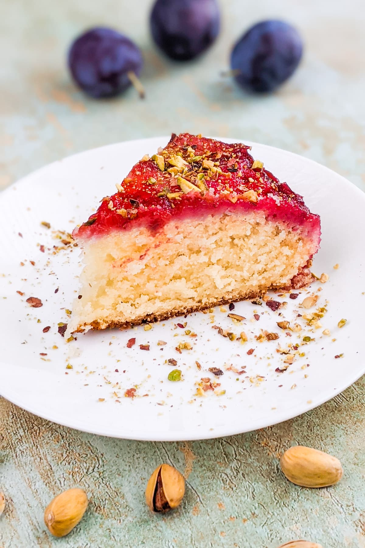 Slice of yogurt plum cake with crushed pistachios on a white plate.