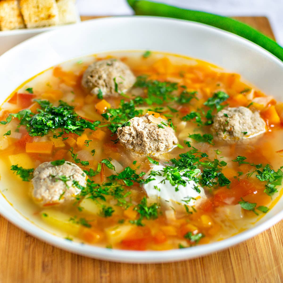 Soup with meatballs and vegetables in a white plate on a wooden cutting board.
