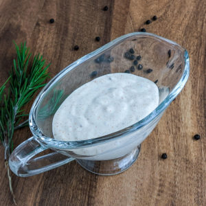 Glass gravy boat with white sauce, rosemary, and black pepper on a wooden table.