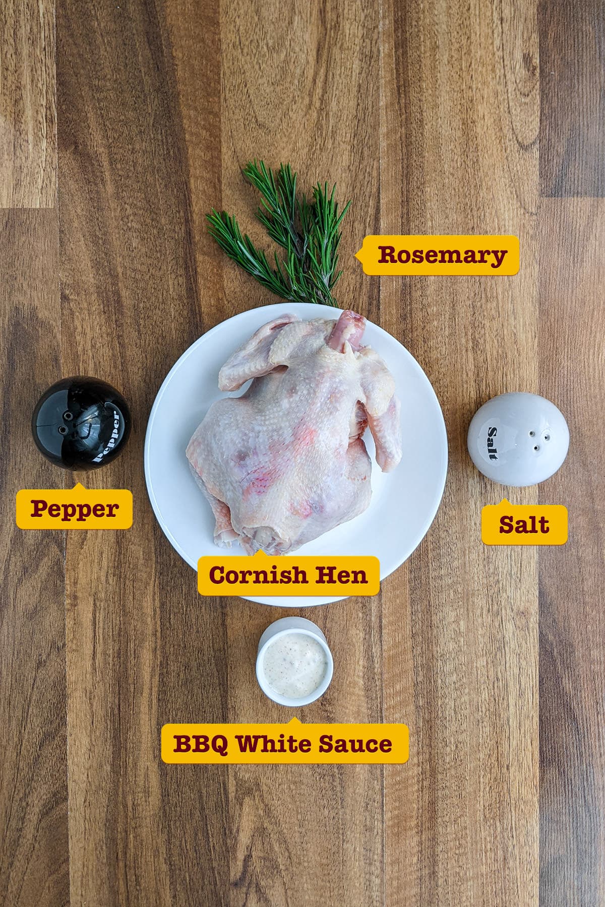 Raw whole cornish hen with salt, pepper and rosemary on a wooden table.