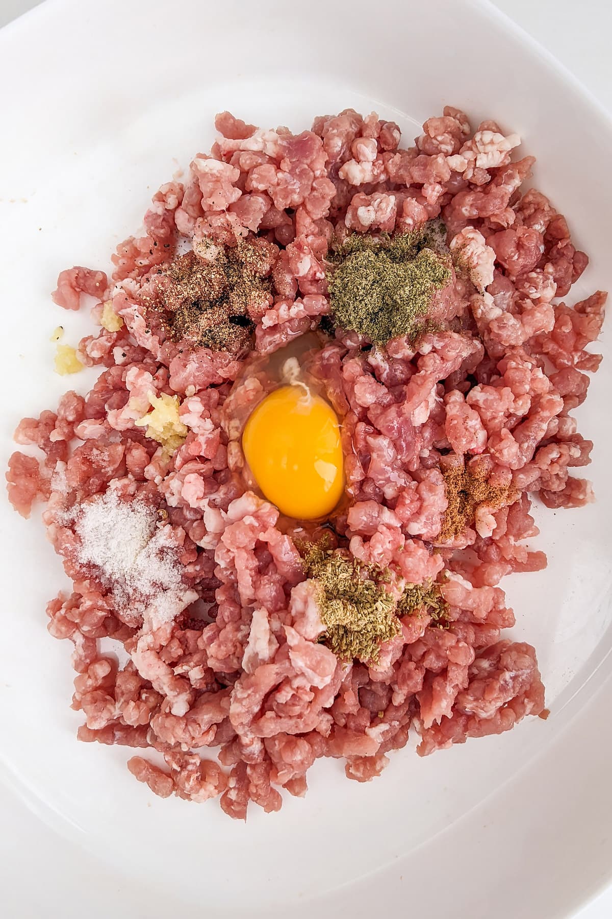 Ground meat pork with one raw egg, and spices in a white big plate.