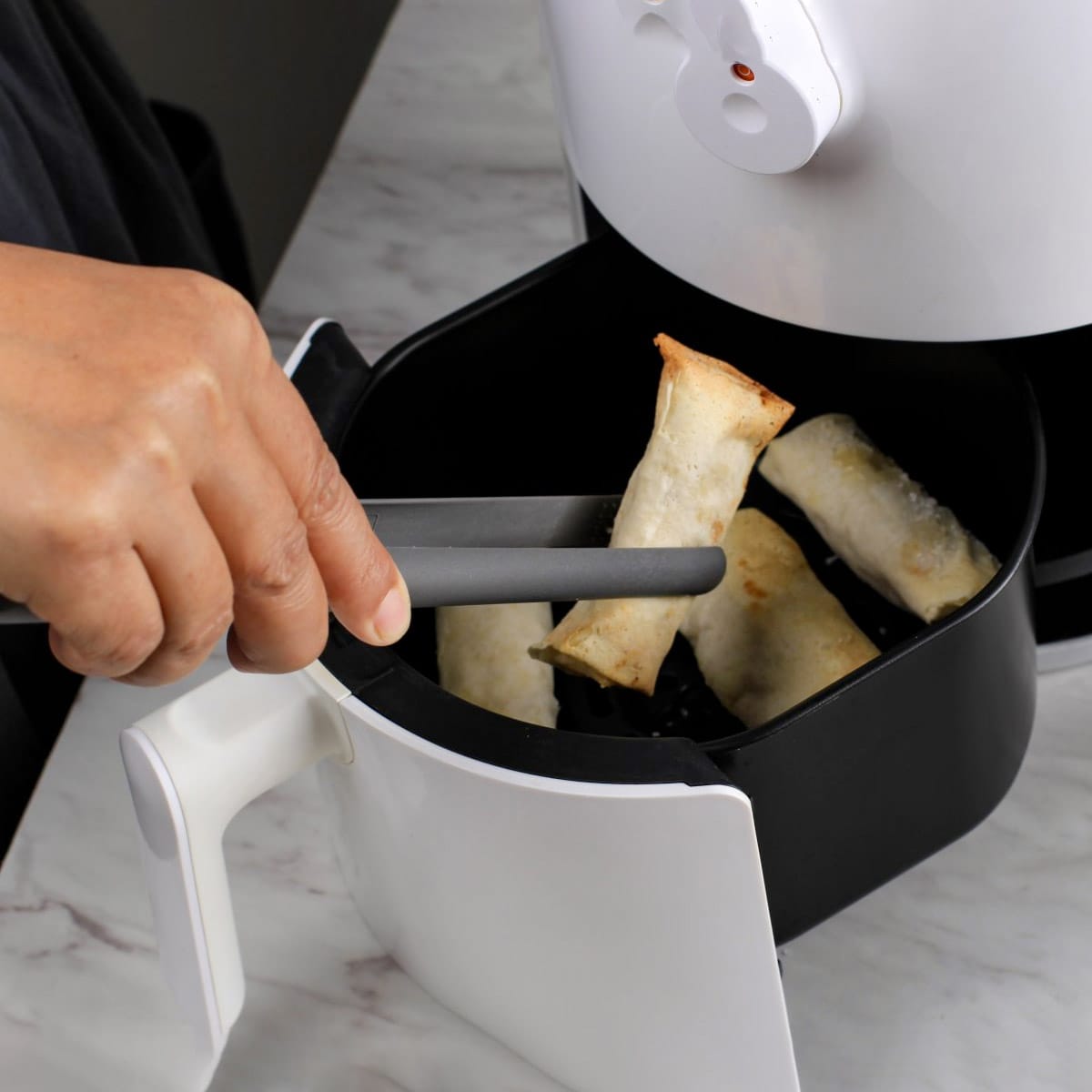Hand with kitchen tongs flipping spring rolls in air fryer.