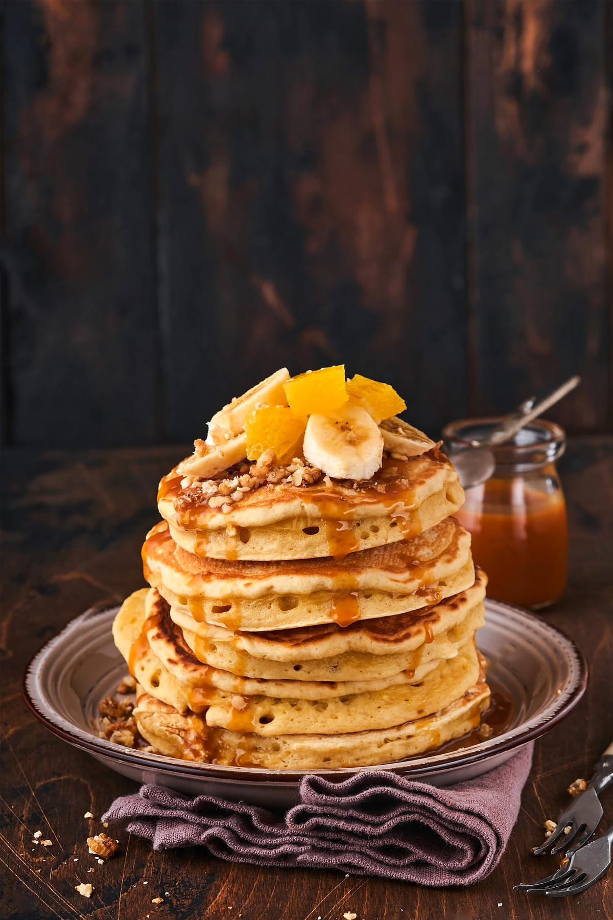 Close look of pancakes with honey, banana slices and orange on a dark wooden background.