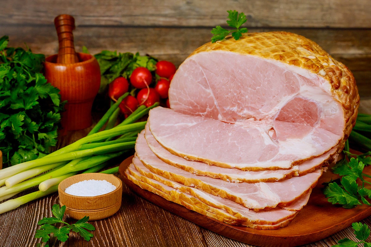 Close look of sliced honey-baked ham in a cutting board with vegetables.