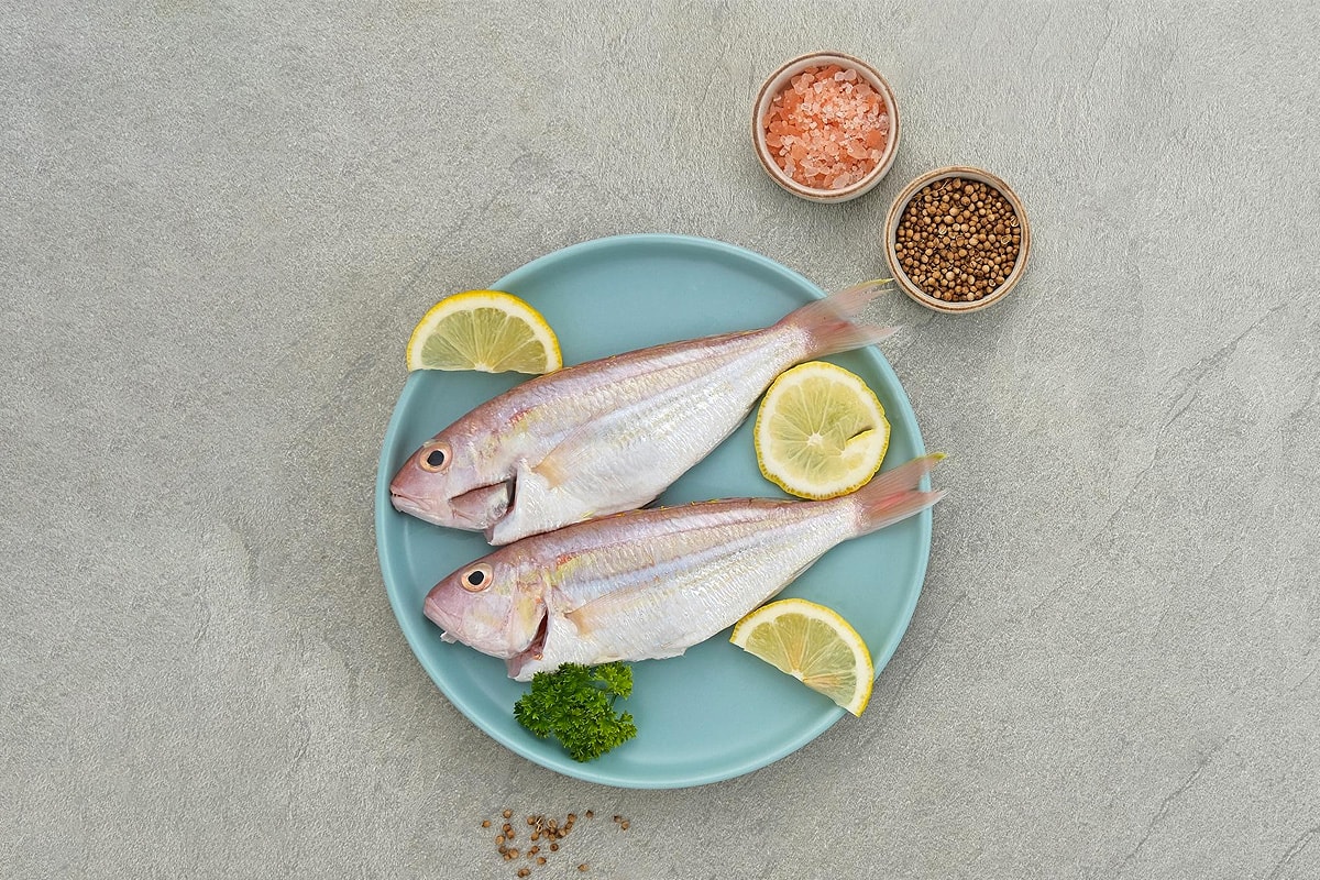 Two cleaned croaker fishes on a blue plate with lemon slices, black pepper and salt.
