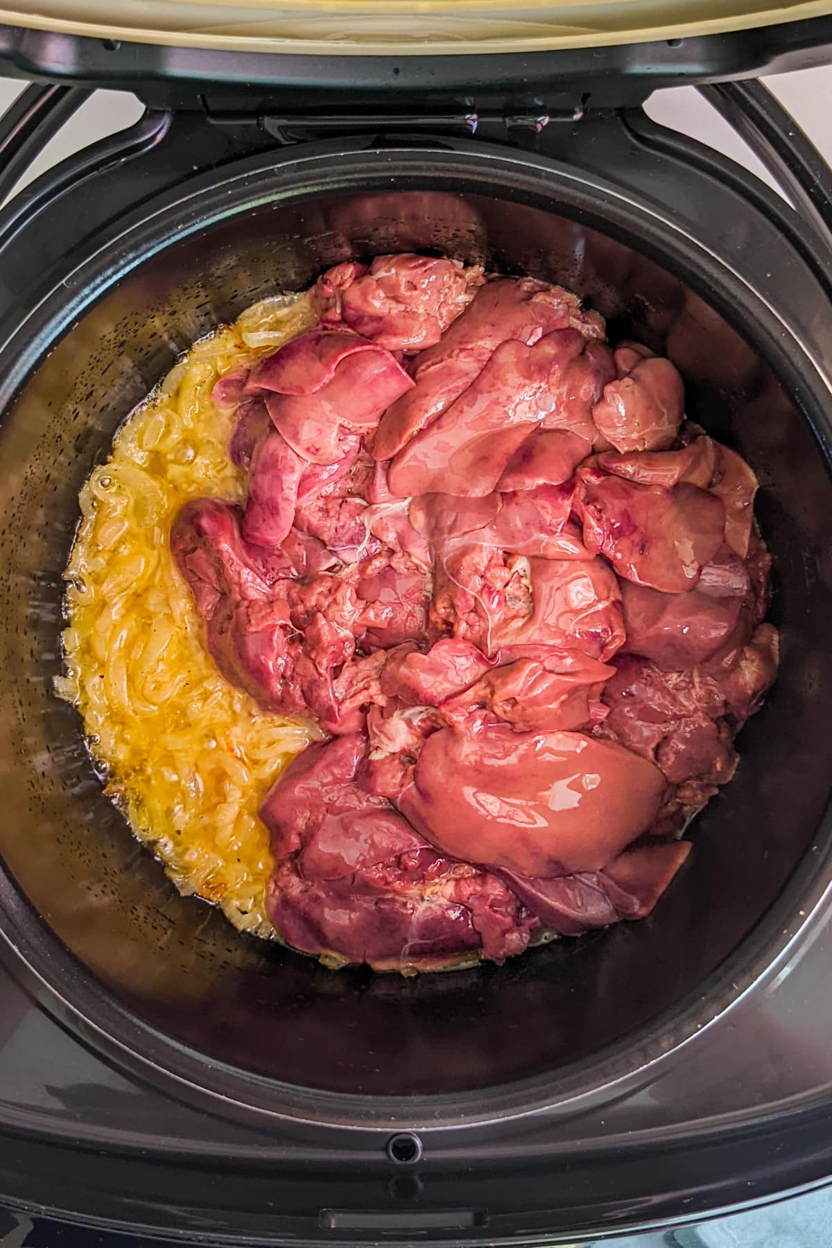Top view of a slow cooker with caramelized onions and raw livers.