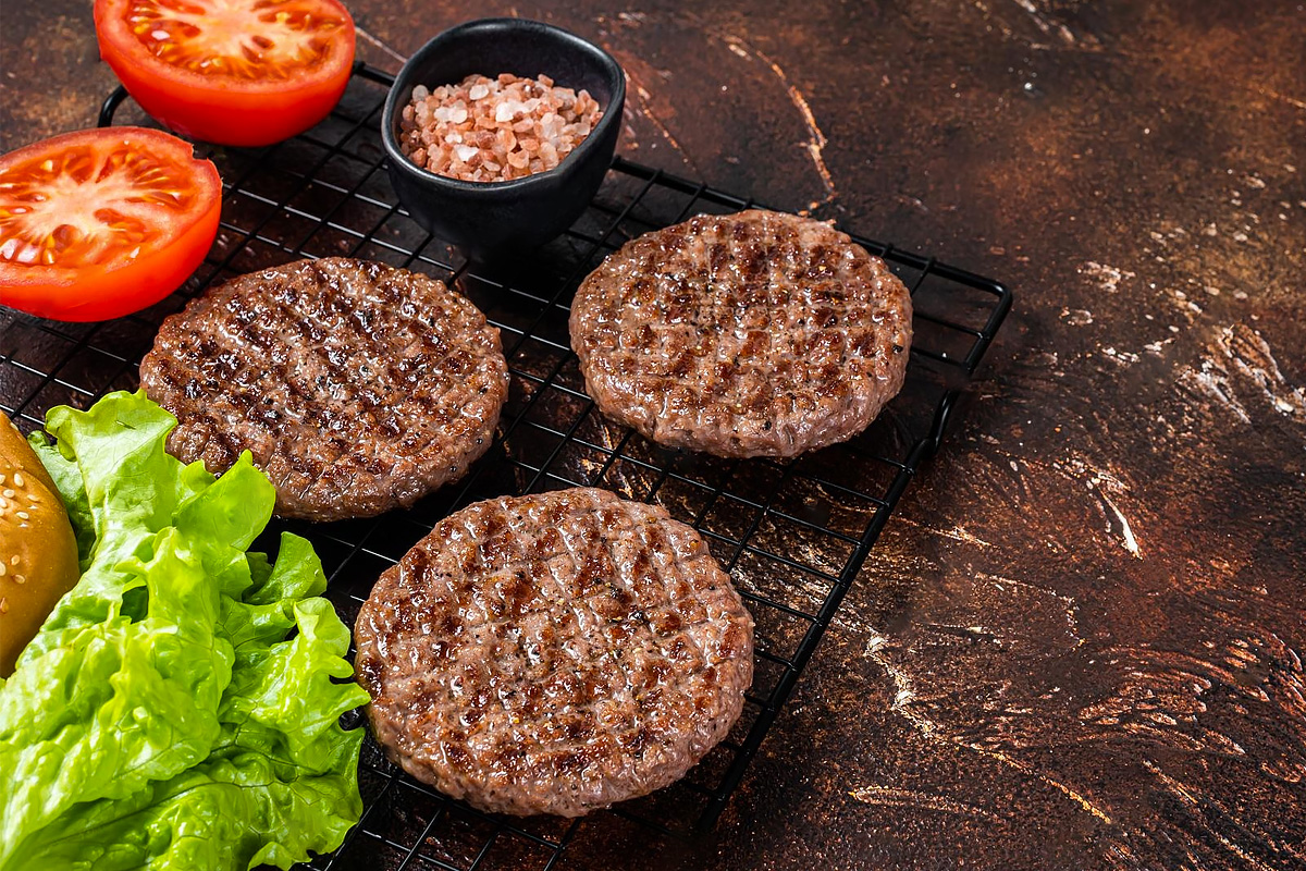 3 burger patties near a salad leave and two halves of a fresh tomato.