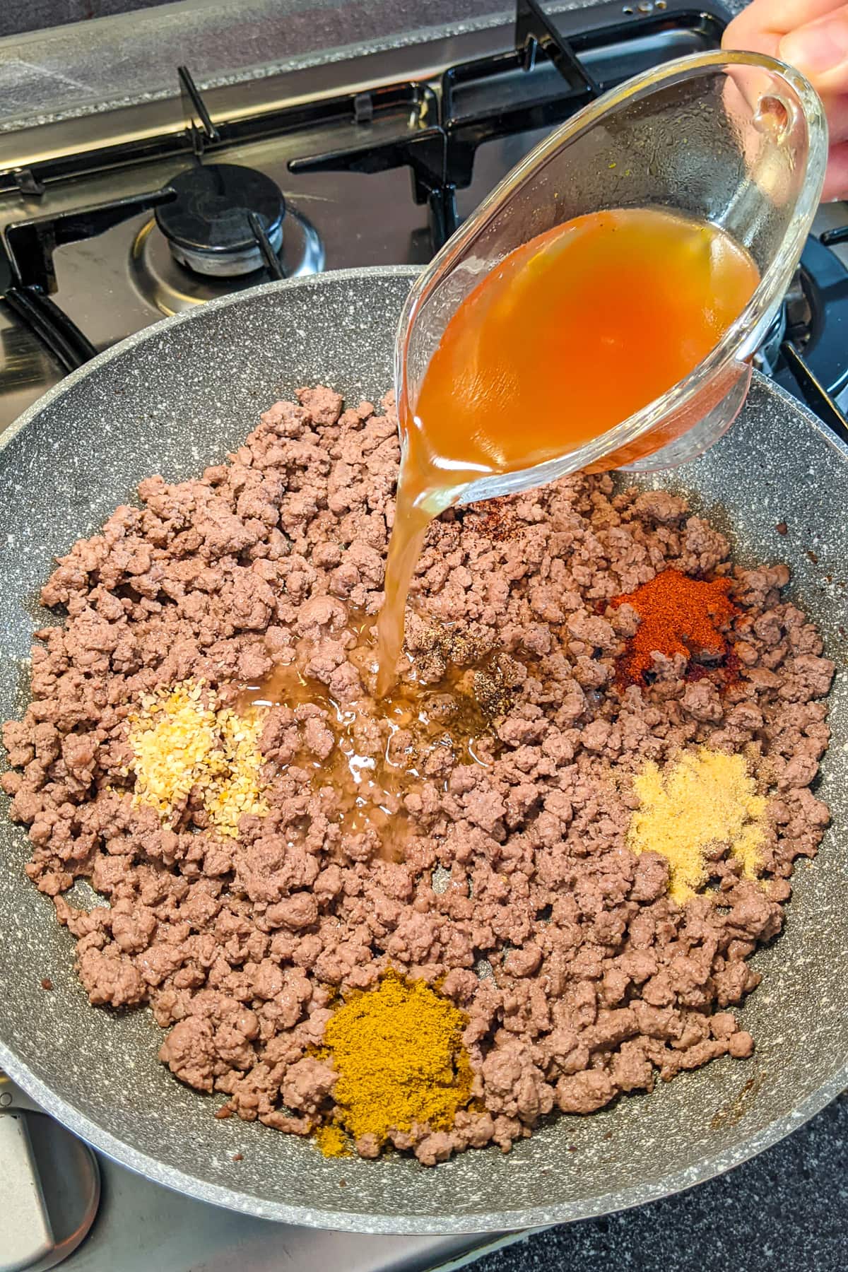 Pouring broth in a frying pan with ground beef with spices.