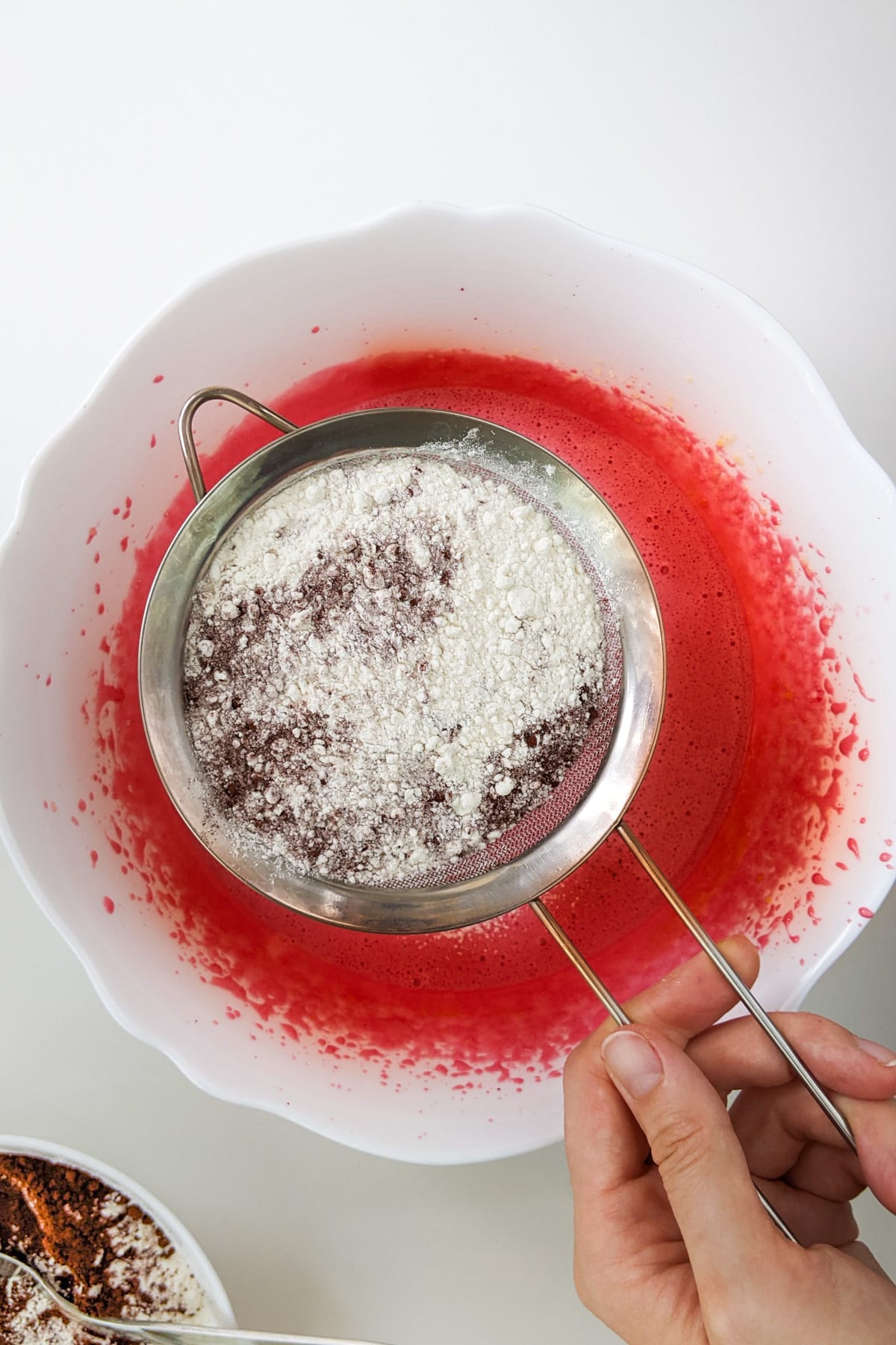 Woman hand holding a sieve with all-purpose flour over red cream.