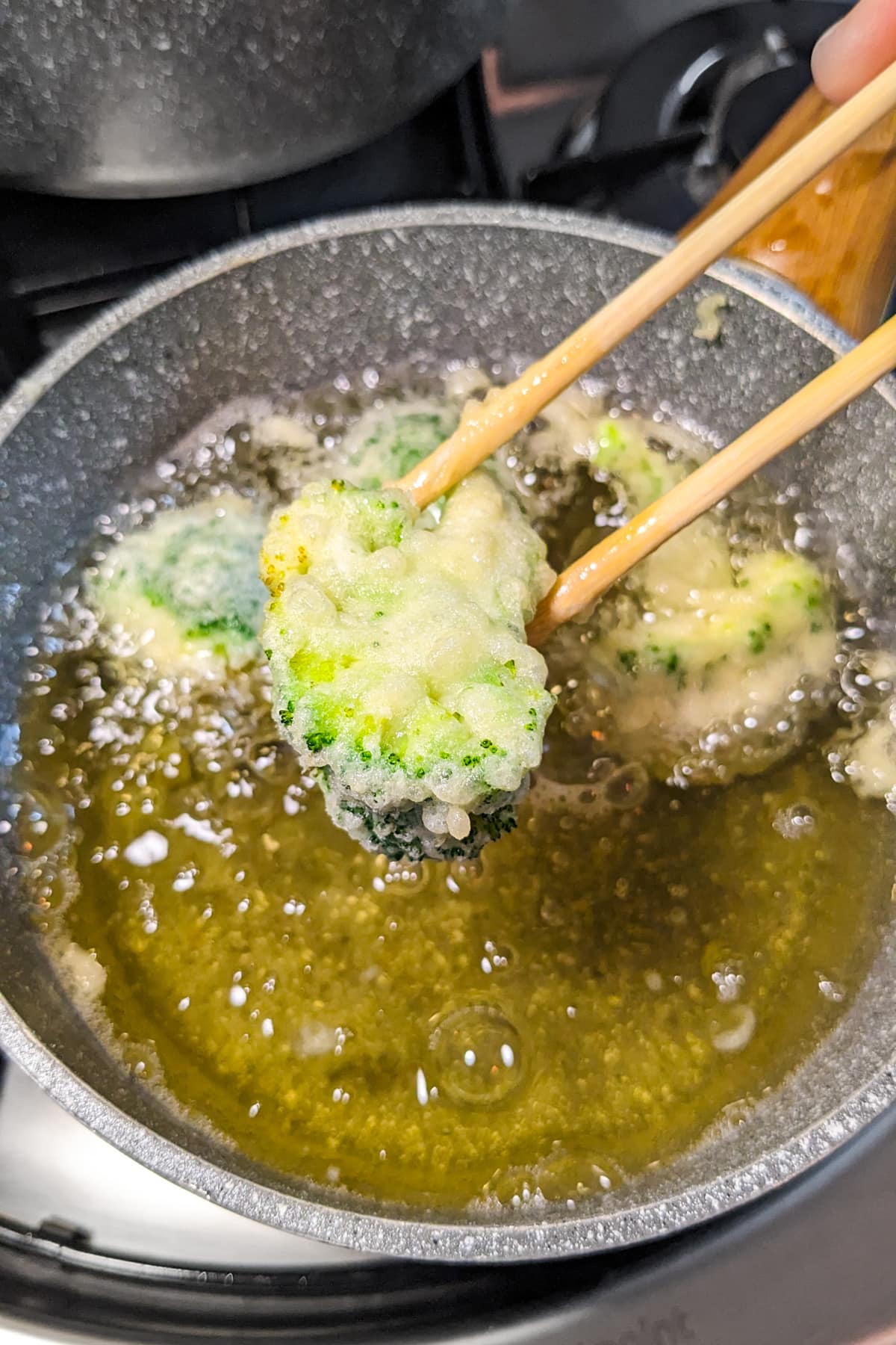 Two wooden sticks holding tempura broccoli over a pan with hot oil.