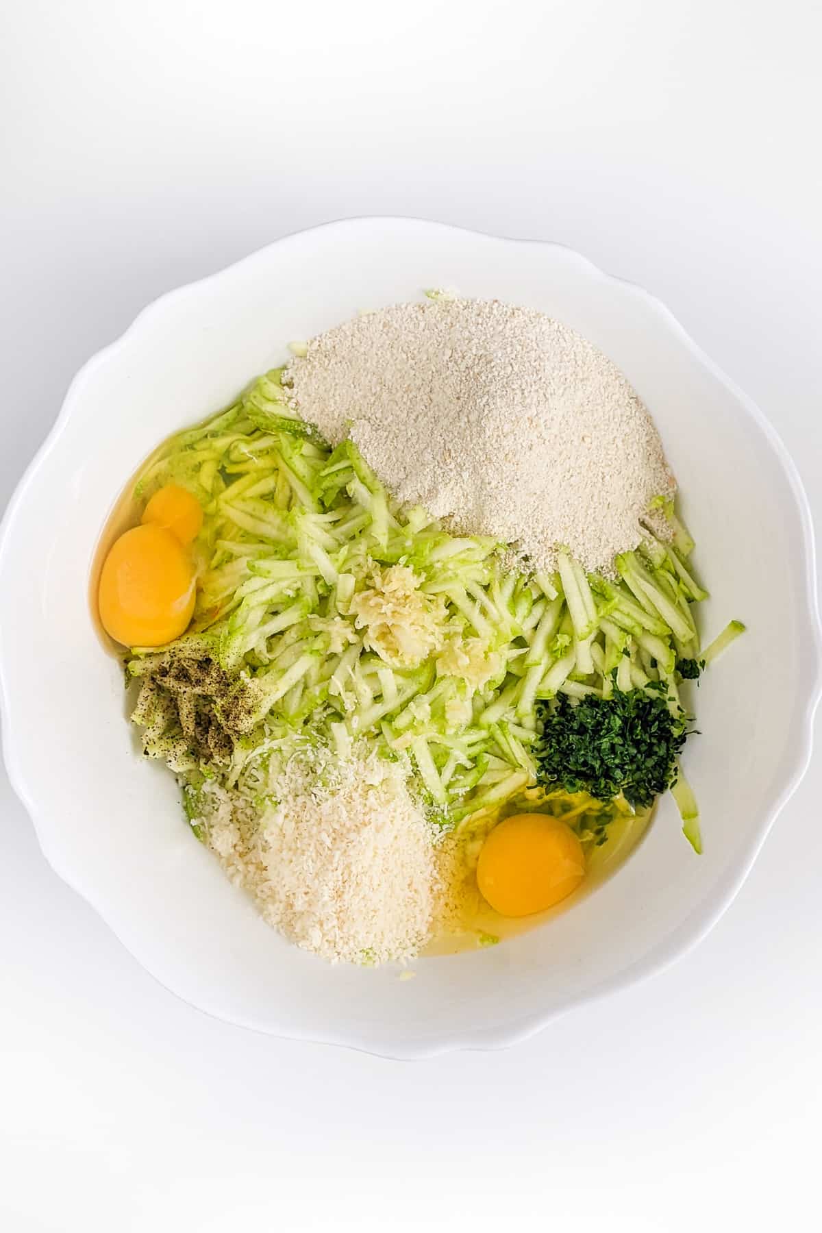 Deep bowl with shredded zucchini, parmesan, breadcrumbs, raw eggs and parsley.