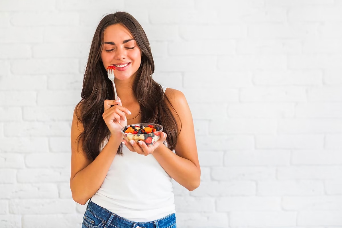 A girl smiling while eating a fruit breakfast on a white background.