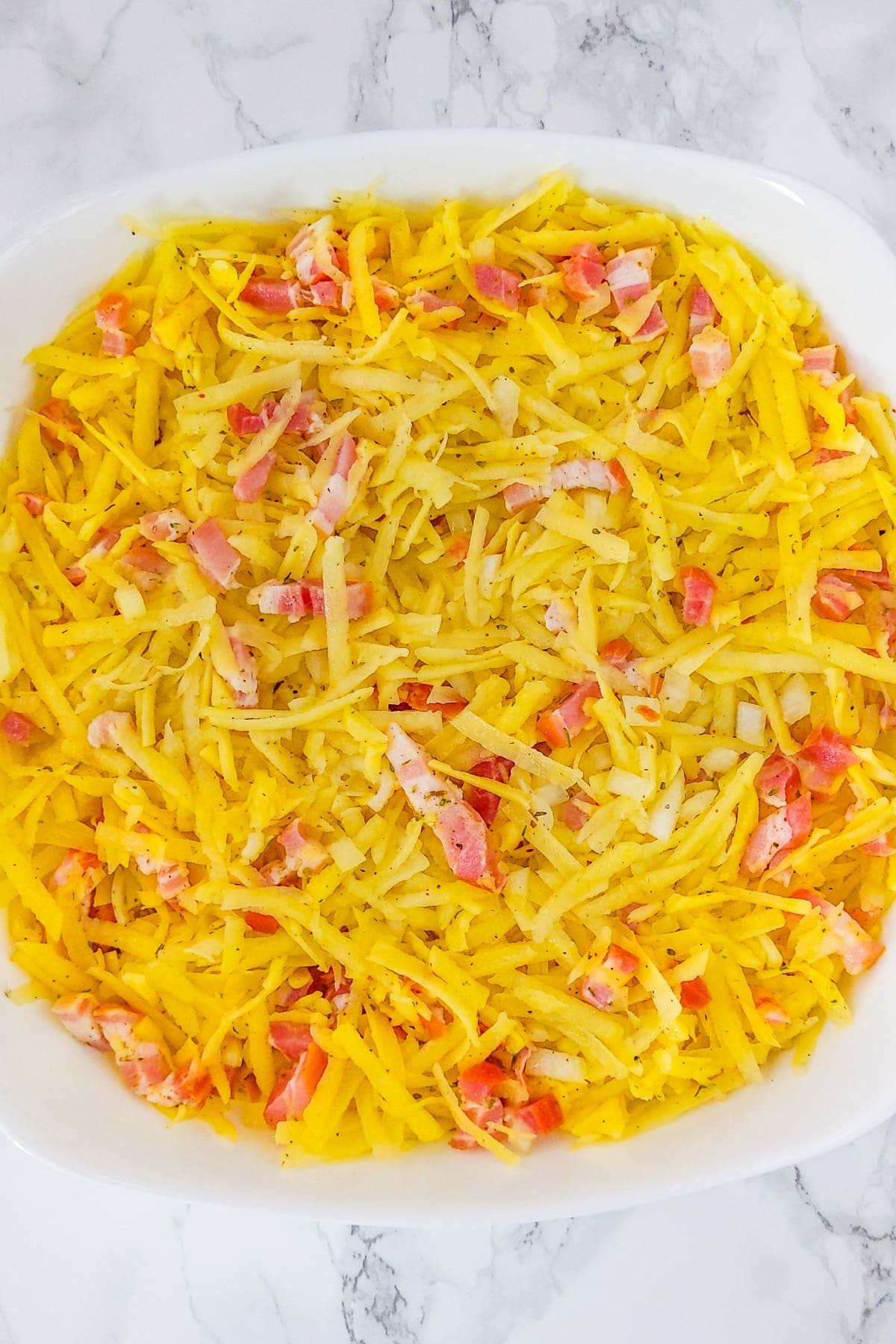 Top view of a white casserole with shredded potatoes, cheddar and sliced bacon.