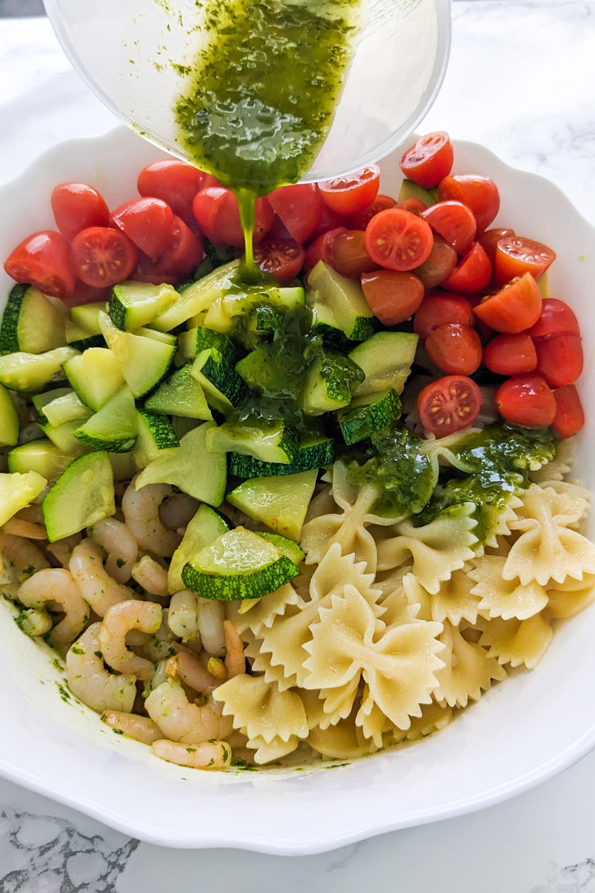 Pouring pesto sauce over cucumbers, cherry tomatoes, pasta and shrimp.