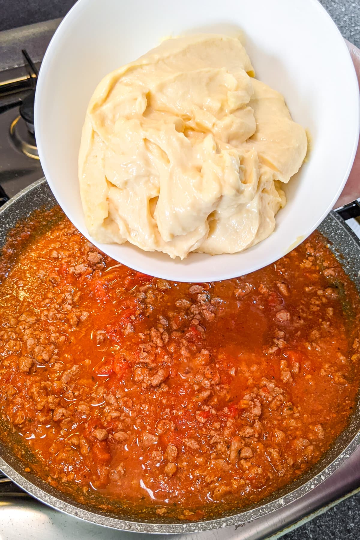 Pouring cheese sauce over fried ground beef in tomato sauce.
