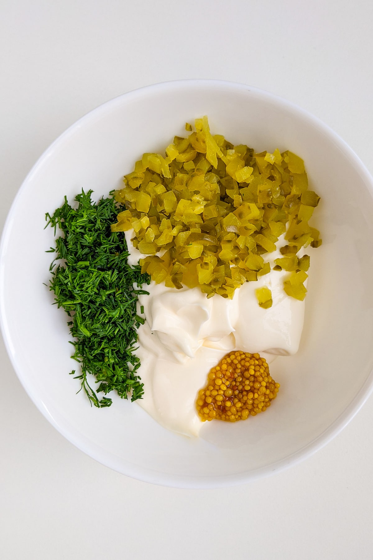 Chopped dill, whole mustard, mayo and pickles in a white plate.
