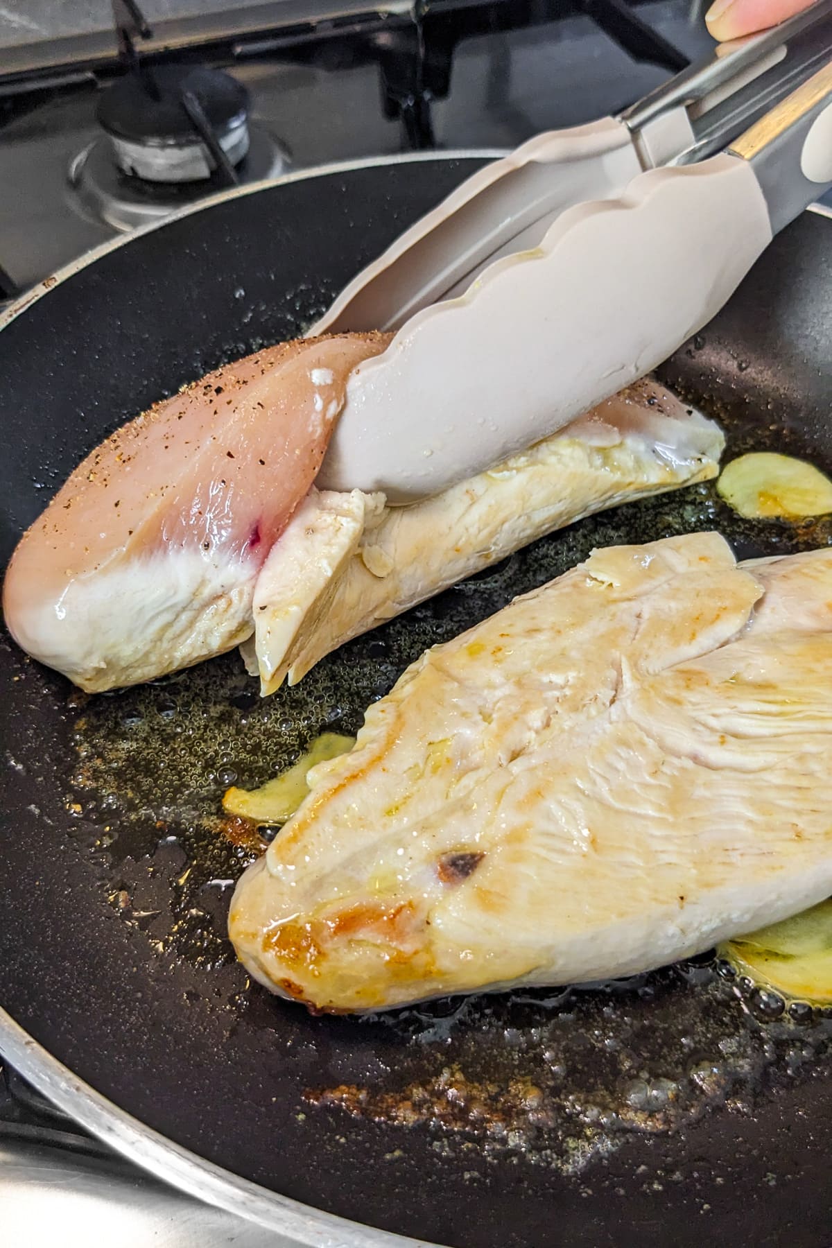Silicone kitchen tongs holding a chicken breast fillet in a frying pan.