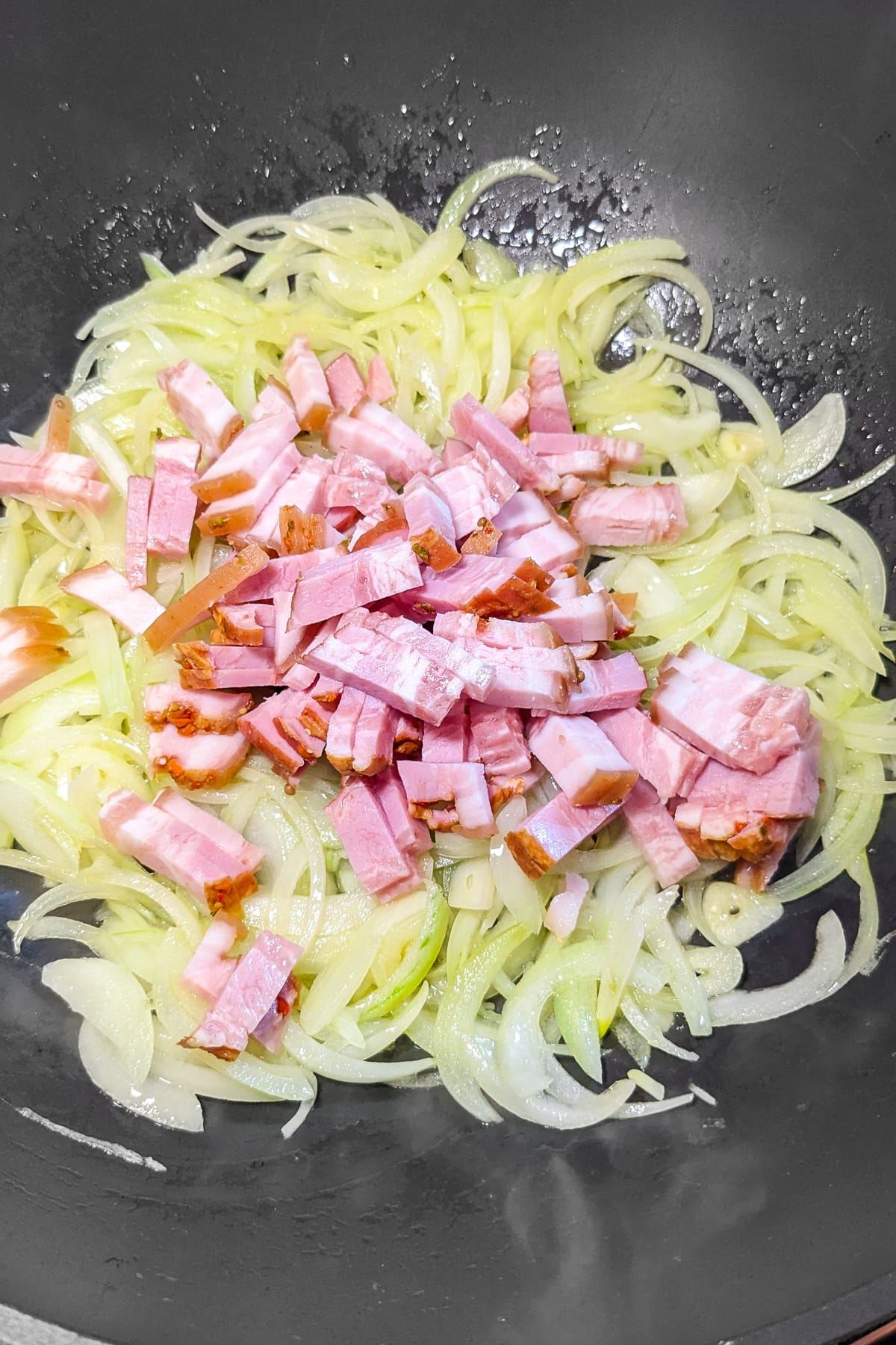 Adding bacon over caramelized onions and garlic cloves in a wok.