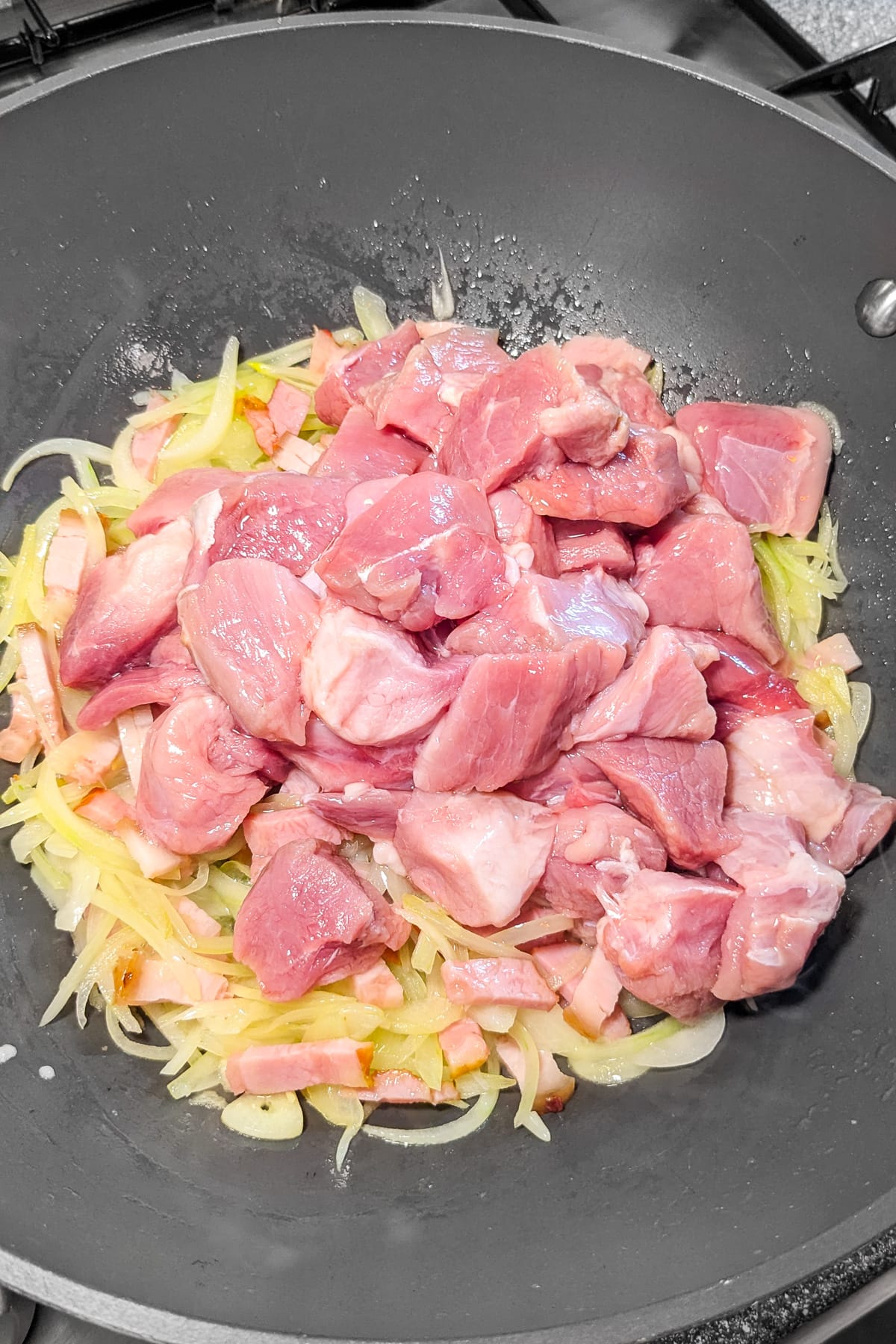 Raw pork meat over a mix of sliced onions and bacon in a wok.