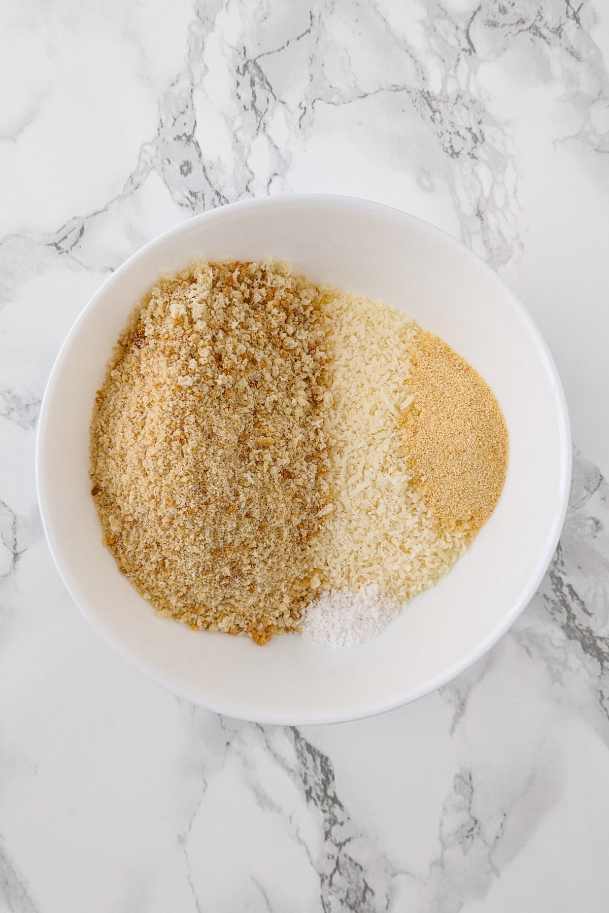 White plate with breadcrumbs, parmesan cheese, and spices on a marble table.