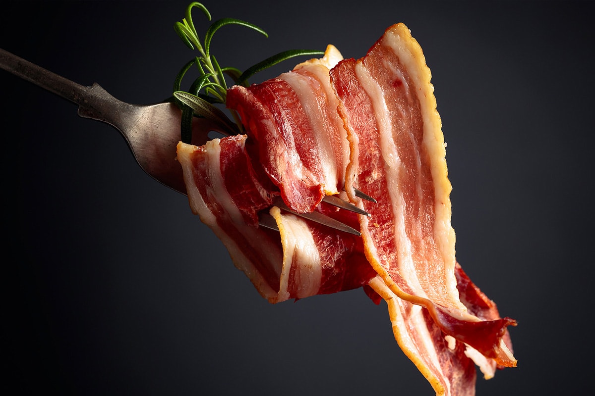 Close look of a fork holding a slice of fried bacon on a black background.