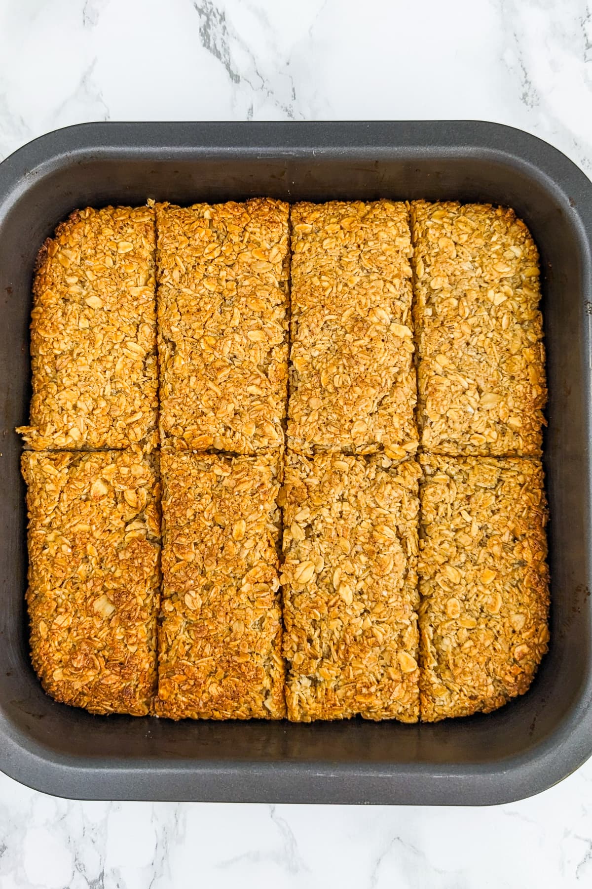 Sliced baked flapjacks in a small square baking tray on a white marble table.