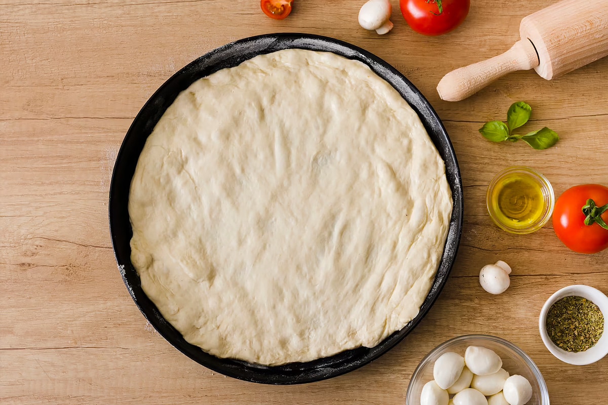 Pizza dough in a baking pan near mozzarella, tomatoes, mushrooms, and olive oil.