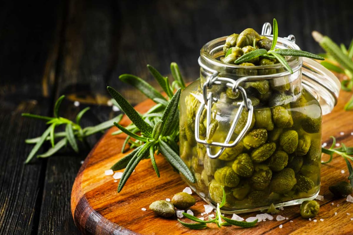 A jar with salted capers and rosemary on a wooden cutting board.