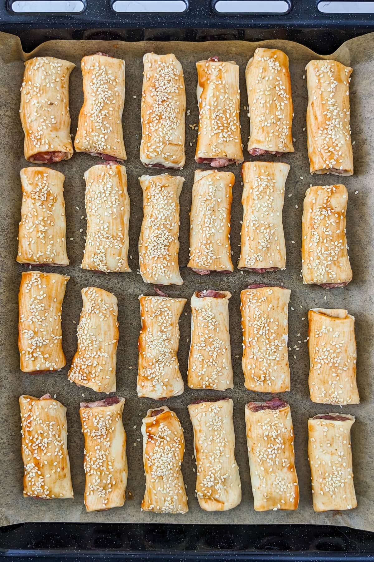 Tray bake full with sesame sausage rolls on a parchment paper.