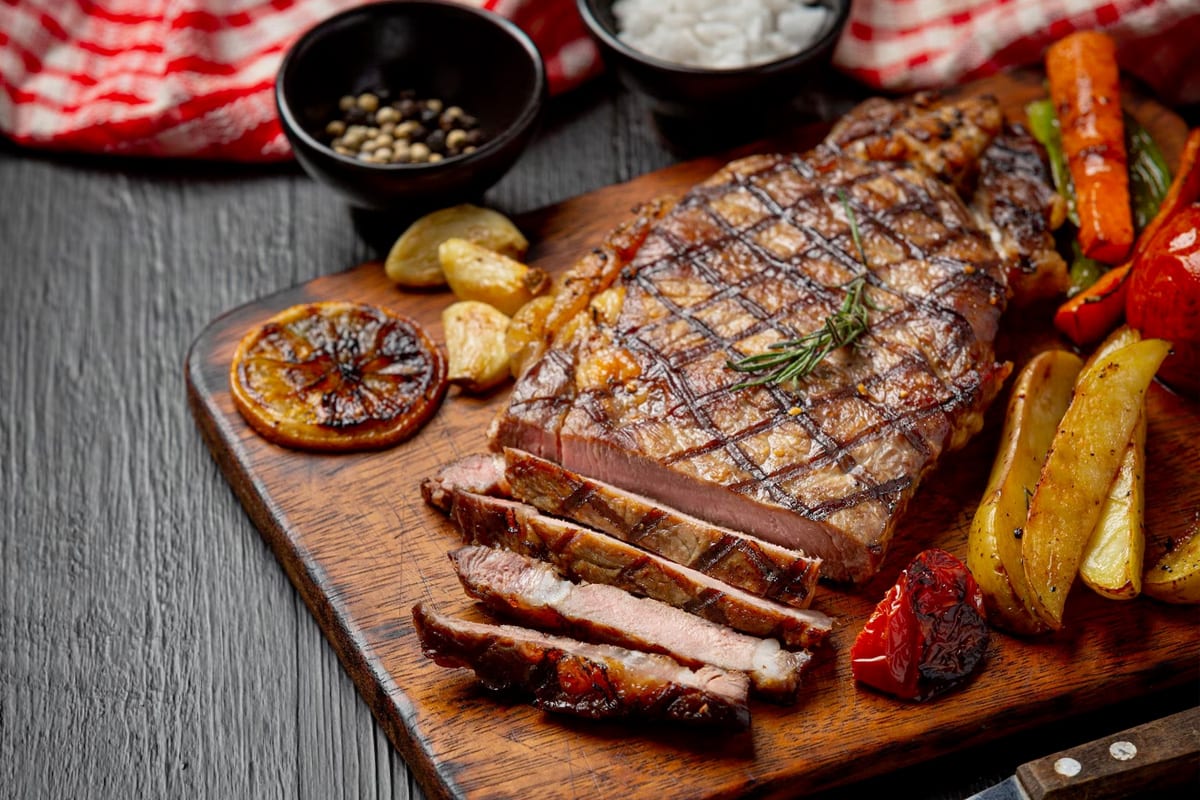 A wooden board with a steak, roasted potatoes, carrots, tomatoes and garlic cloves.