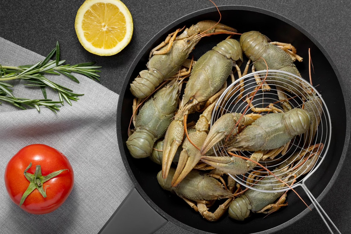 A black frying pan with raw crawfish near a fresh tomato, a slice of lemon and rosemary.