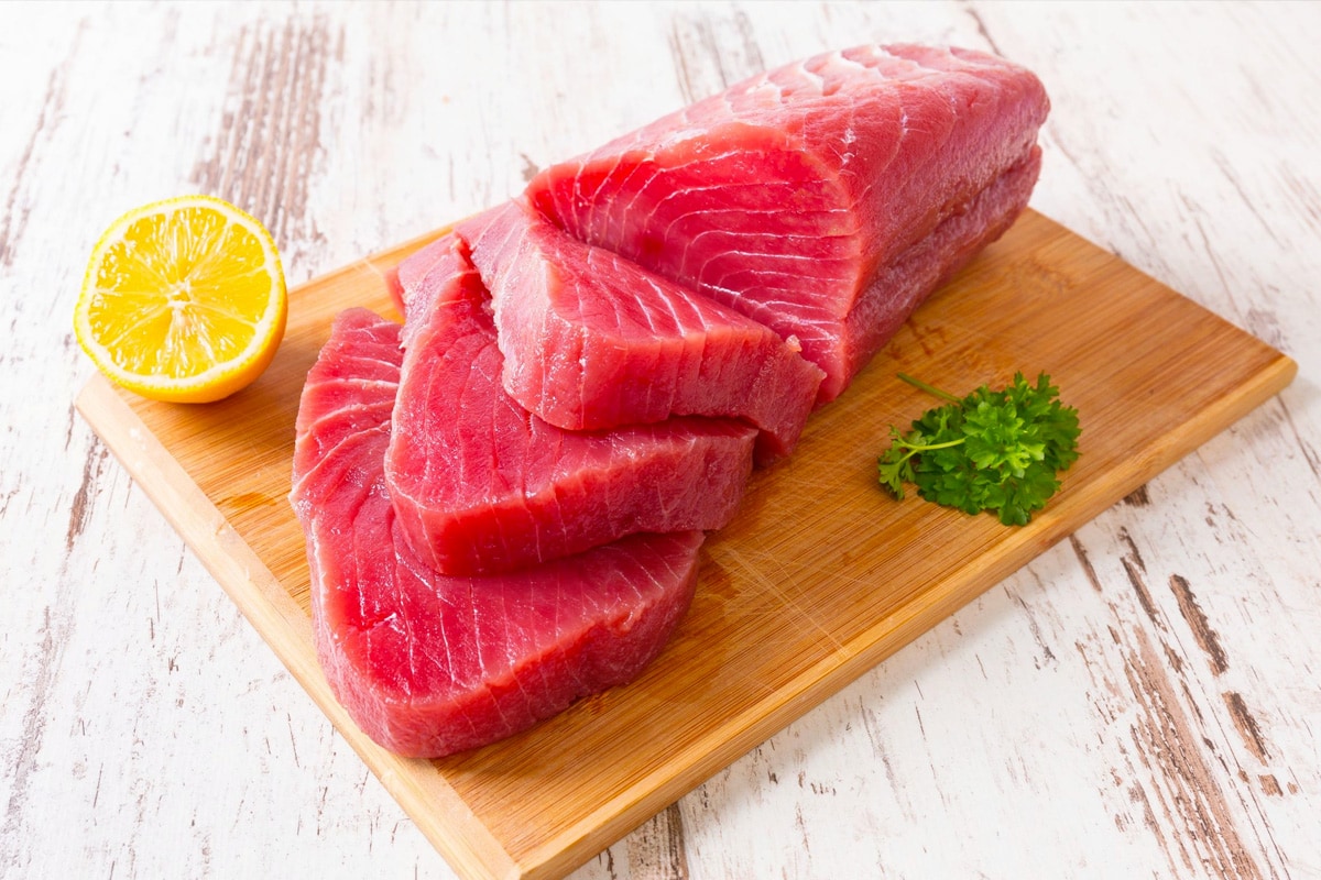 A wooden cutting board with a few slices of tuna meat near parsley and fresh lemon.