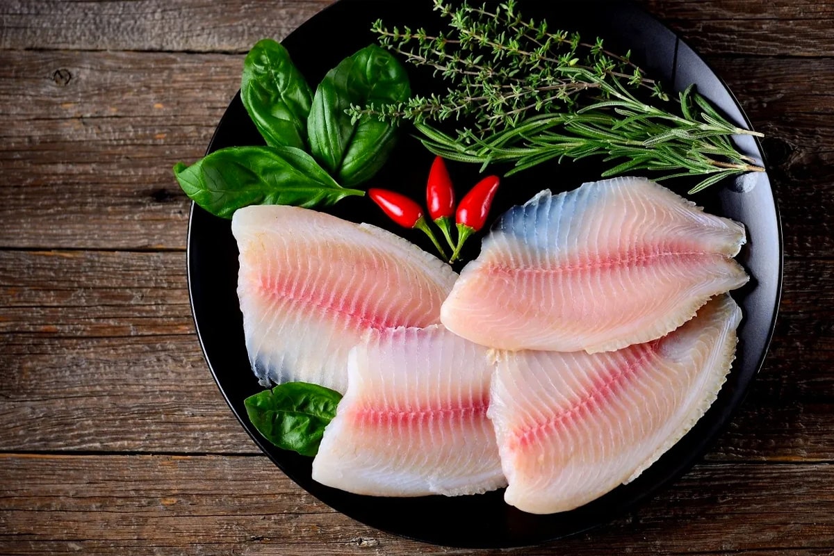 Top view of raw tilapia with aromatic herbs in a black plate on a wooden table.