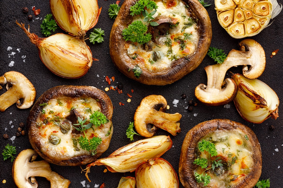 Top view of stuffed mushrooms near some roasted onions and aromatic herbs.