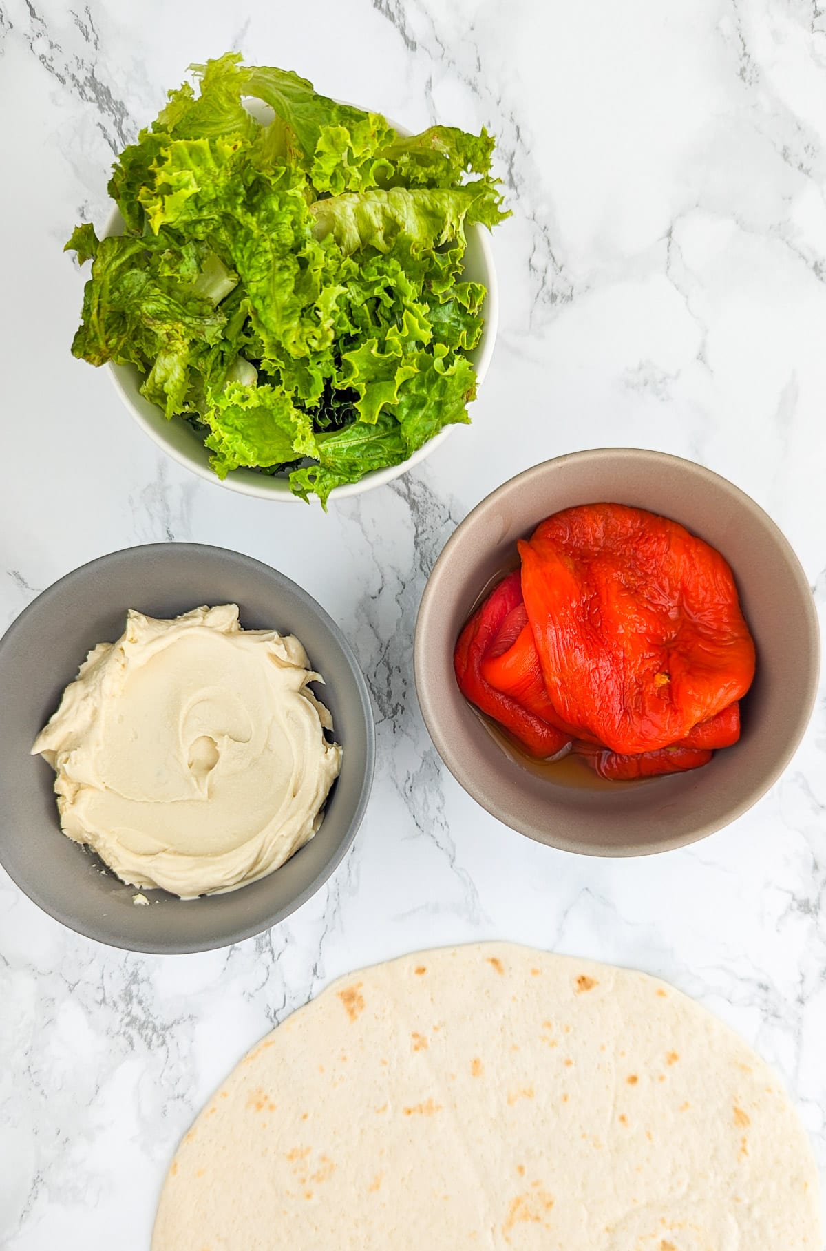Top view of a bowl with salad, one with cream and one with roasted bell peppers near a slice of pita.