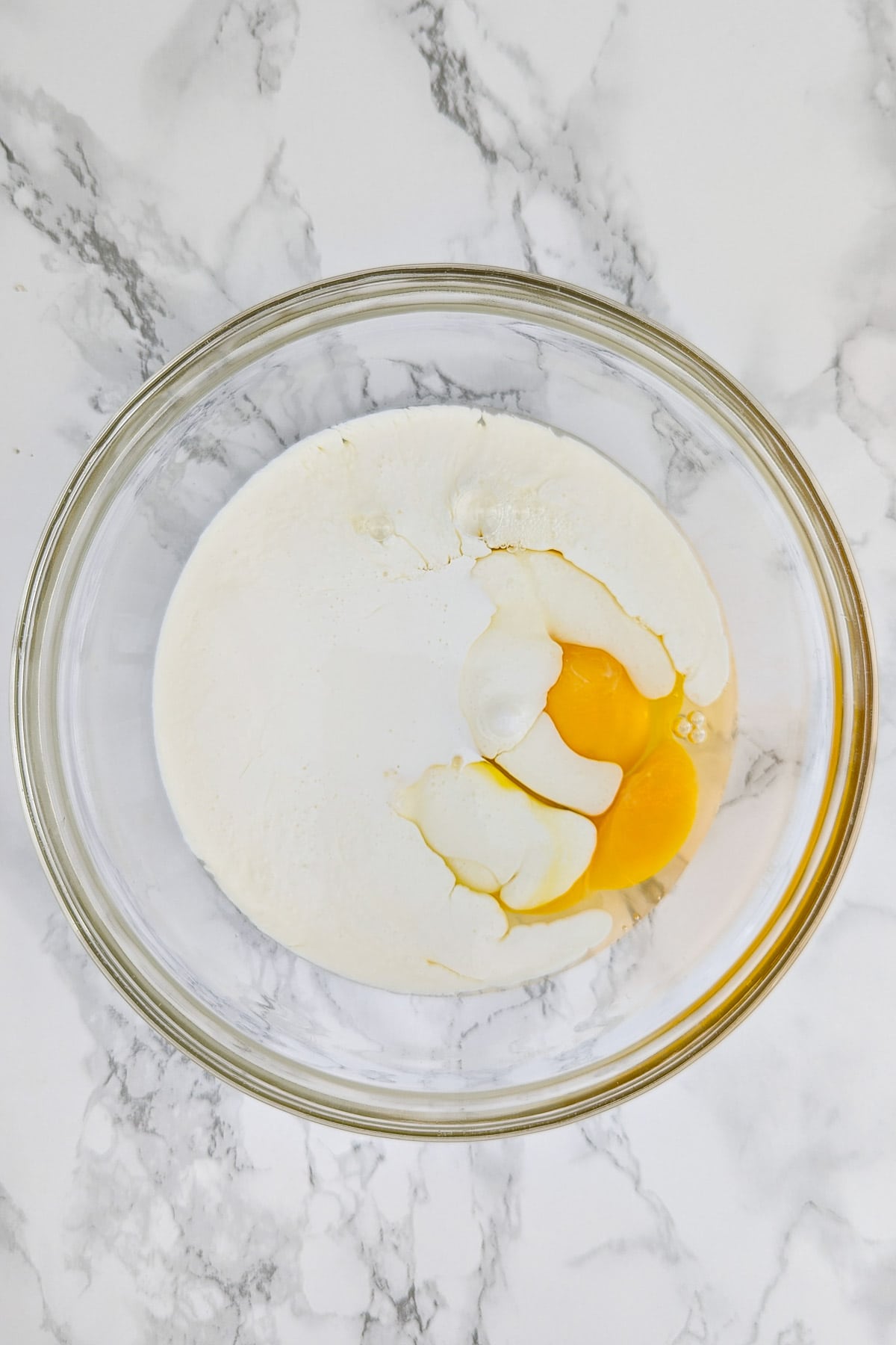 Two raw eggs and cream in a transparent bowl on a marble table.