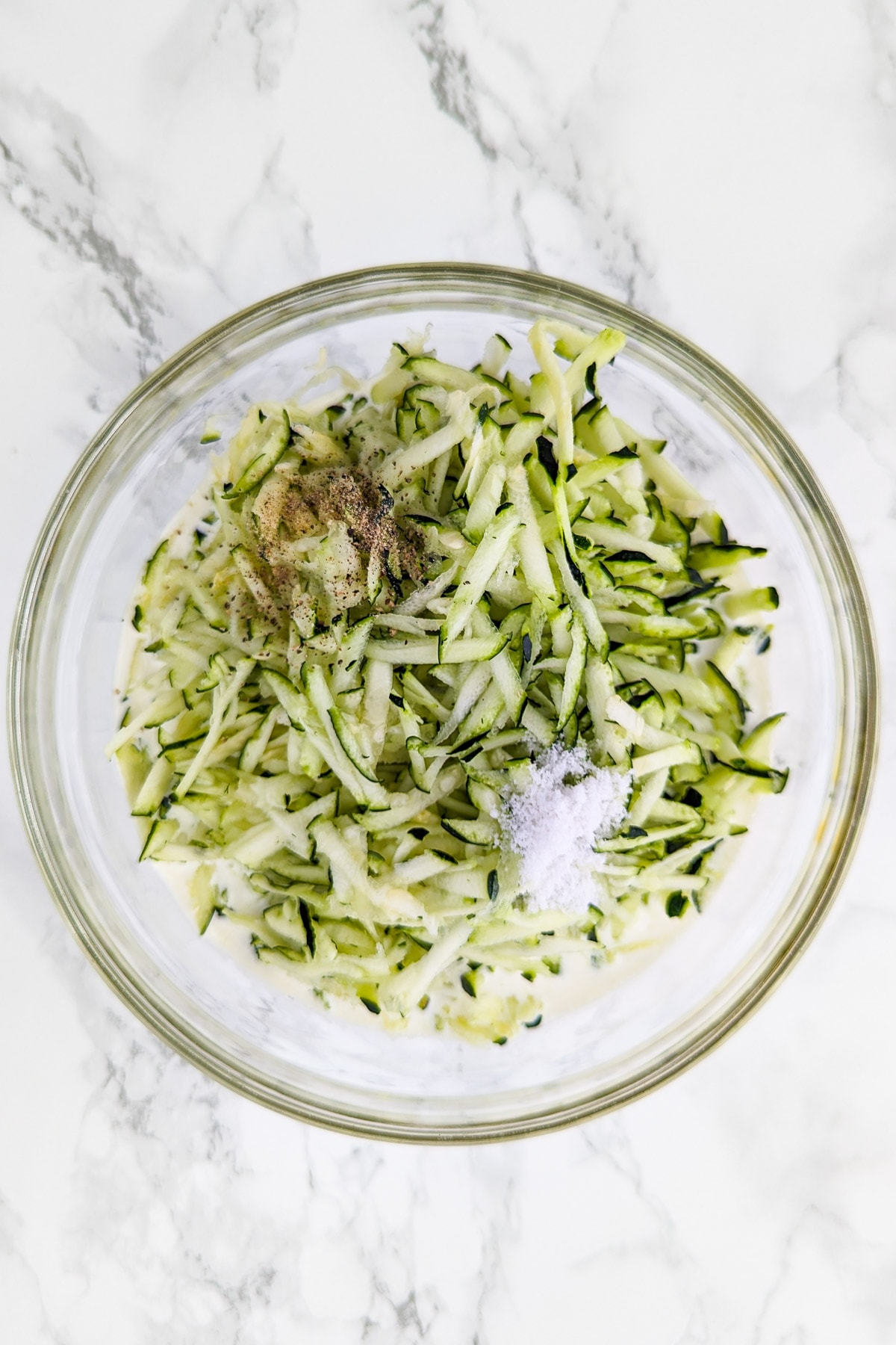 Shredded zucchini with salt and black pepper in a white transparent bowl.