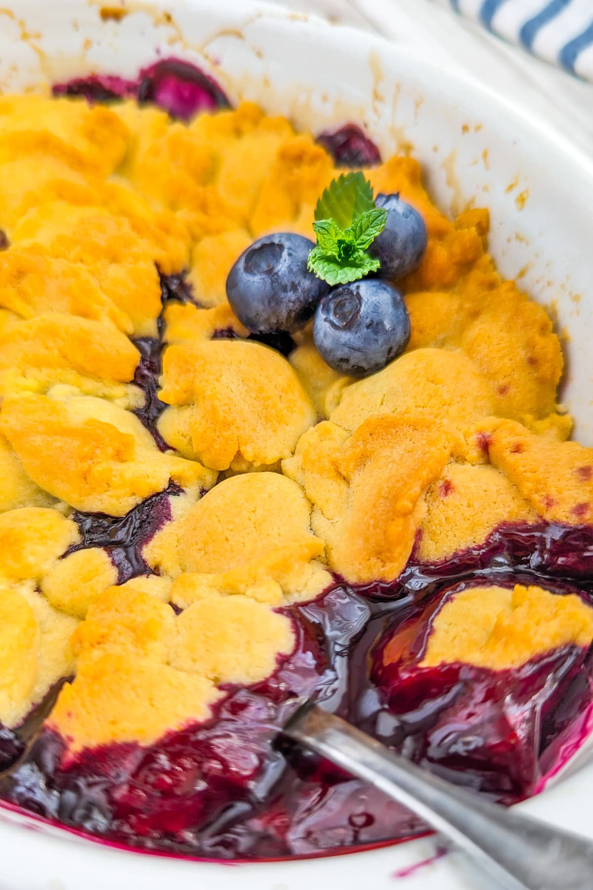 Close look of a blueberry cobbler decorated with 3 blueberries, mint and a spoon in it.