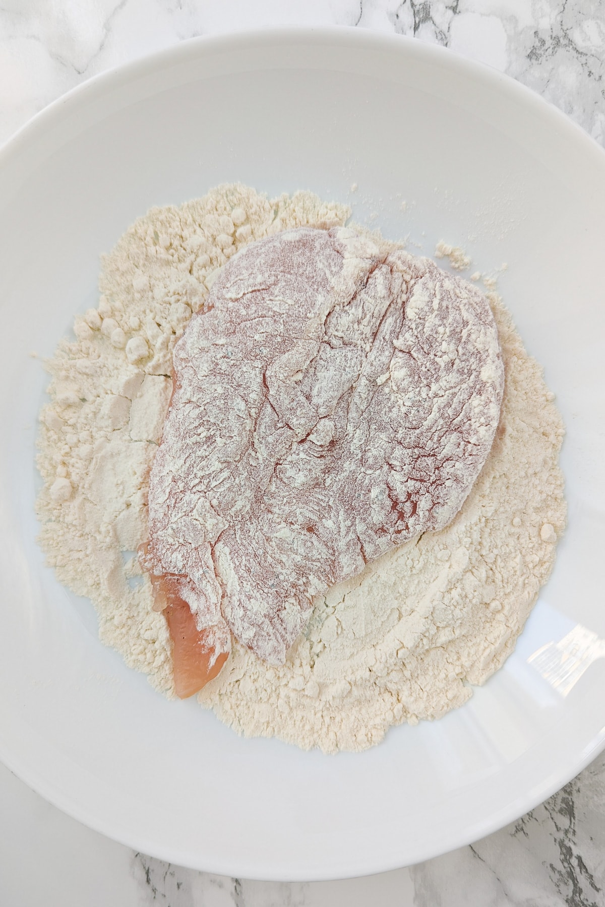 Top view of white bowl with chicken breast covered in all-purpose flour.