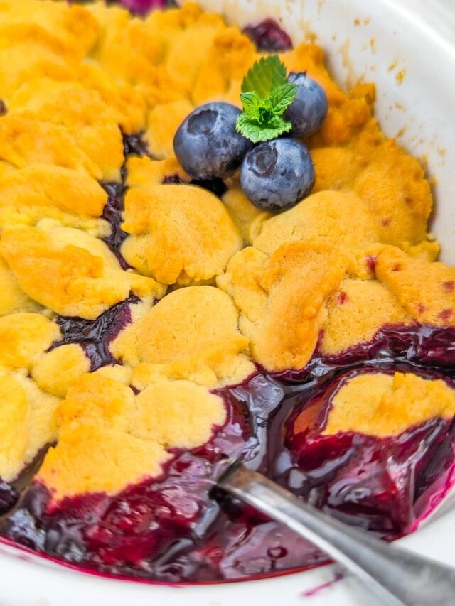 Close look of a blueberry cobbler decorated with 3 blueberries, mint and a spoon in it.