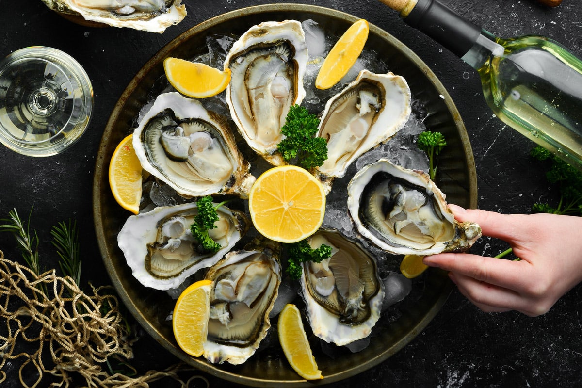 Top view of a woman hand holding an oyster over a plate full with oysters and lemon slices.