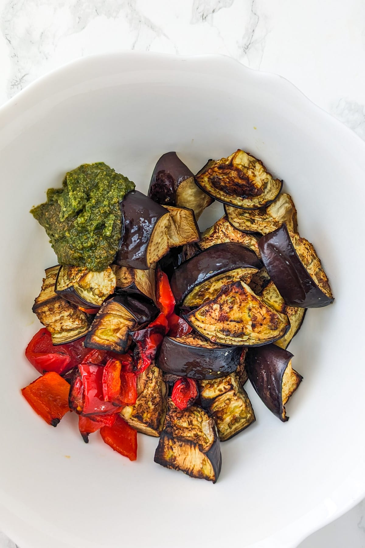 Top view of a white deep bowl with roasted eggplants, bell peppers and pesto sauce.
