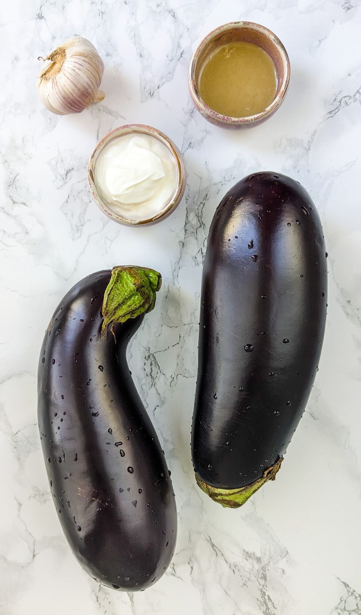 Two eggplants, yogurt and garlic on a white marble table.