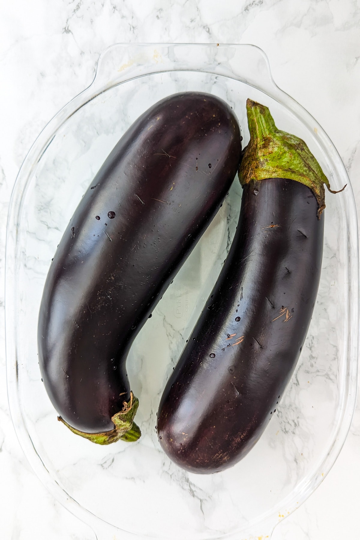 Top view of 2 raw eggplants sitting in a transparent baking dish.